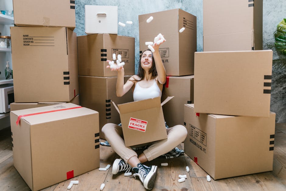 Two women stand inside a room surrounded by stacked cardboard boxes, some sealed with red packing tape. One woman, dressed in a checked shirt and beige trousers, holds a box with a white label, while 