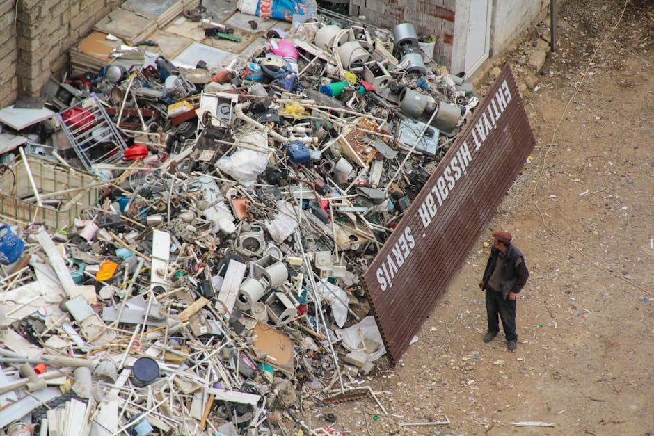 An outdoor view showing a large pile of mixed waste materials, including broken household items, plastic containers, metal parts, and discarded furniture, situated next to a metal fencing panel with w