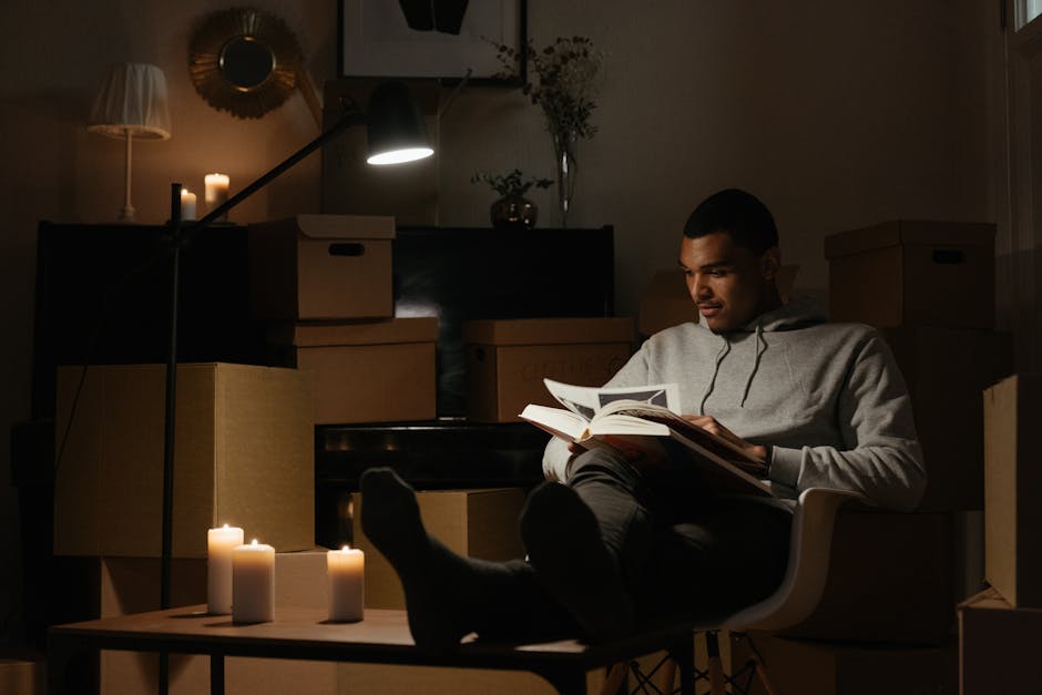 Inside a dimly lit room with a warm, cozy atmosphere, a young man is seated in a modern white chair, reading a book. The room is filled with cardboard boxes of various sizes, some stacked on top of ea