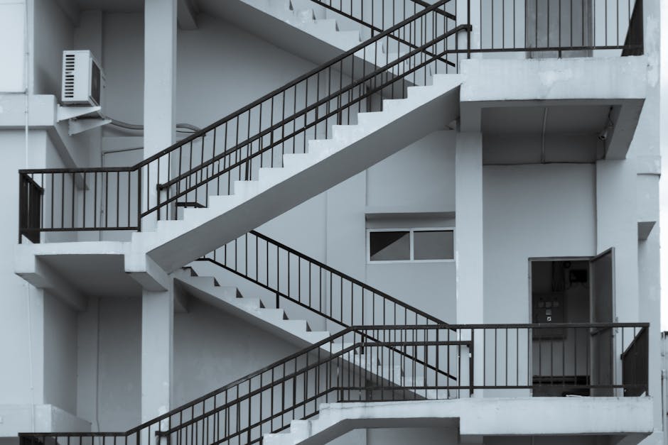 The image displays a modern external staircase with three flights, constructed from metal and glass, attached to the beige concrete facade of a multi-storey residential building. The stairs feature bl